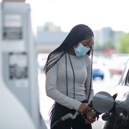 A young woman pumps gas at gas pump. She is wearing a blue medical mask and blue protective gloves.