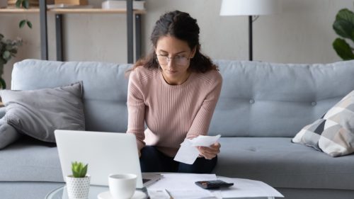 Serious young woman in glasses sit in living room pay bills taxes on laptop online, focused millennial female manage family expenditures expenses, plan budget on computer, make payment on web
