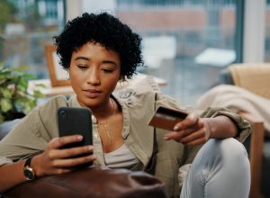 Young woman holding her credit card while using her cellphone at home