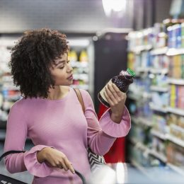 woman in her 20s shopping in a grocery store, carrying a shopping basket. She is reading the ingredient label on a bottle.