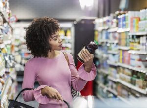 woman in her 20s shopping in a grocery store, carrying a shopping basket. She is reading the ingredient label on a bottle.