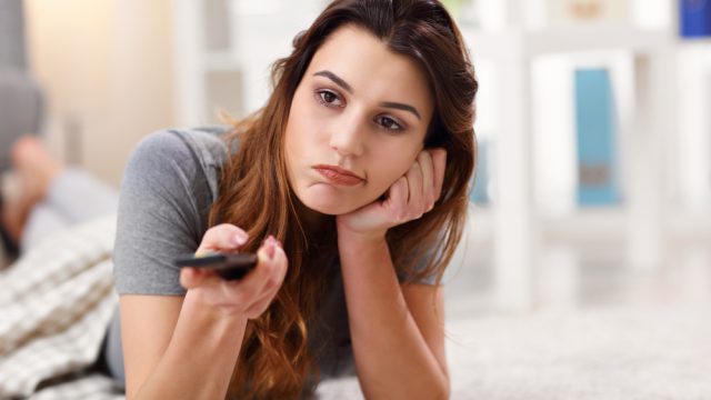 A young woman sitting on the ground watching TV while holding a remote control with a bored look on her face