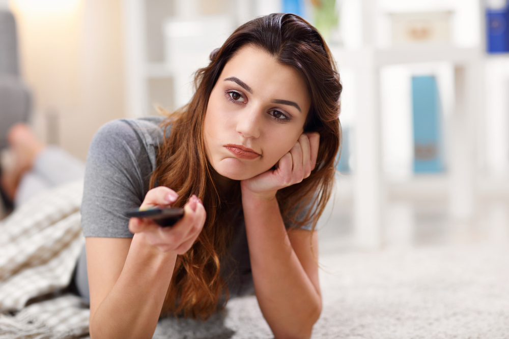 A young woman sitting on the ground watching TV while holding a remote control with a bored look on her face