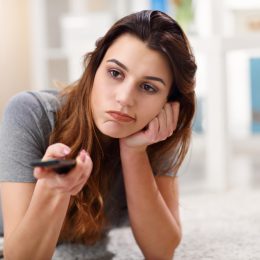 A young woman sitting on the ground watching TV while holding a remote control with a bored look on her face