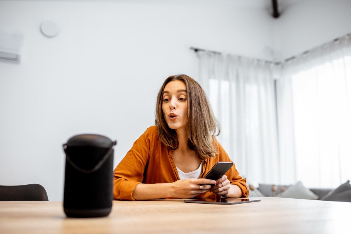 young woman sitting at table holding phone and speaking a command to a smart speaker