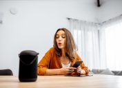 Young cheerful woman controlling home devices with a voice commands, talking to a smart column at home. Concept of smart home and voice command control