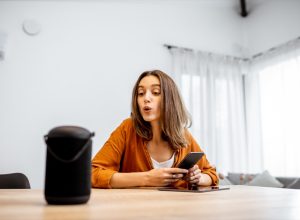 Young cheerful woman controlling home devices with a voice commands, talking to a smart column at home. Concept of smart home and voice command control