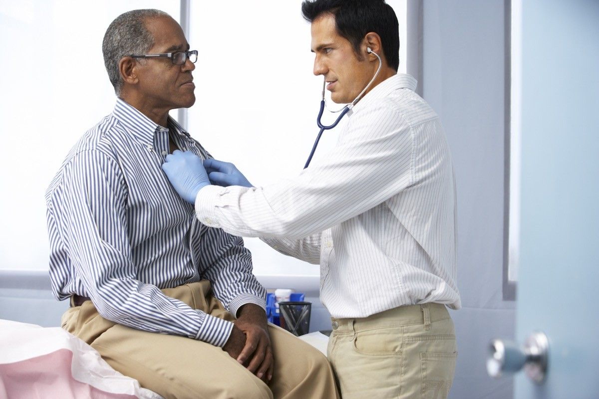 Doctor In Surgery Listening To Male Patient's Chest Using A Stethoscope