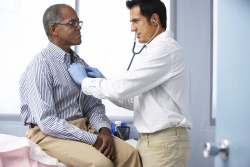 Doctor In Surgery Listening To Male Patient's Chest Using A Stethoscope