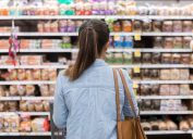 In this rear view, an unrecognizable woman stands with a shopping cart in front of a shelf full of food in the bread aisle of a grocery store.