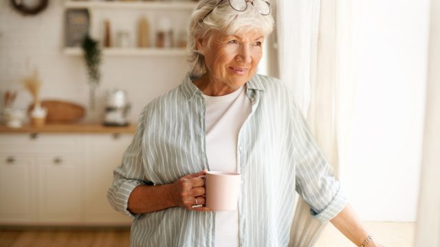 A senior woman standing by the window while holding a mug