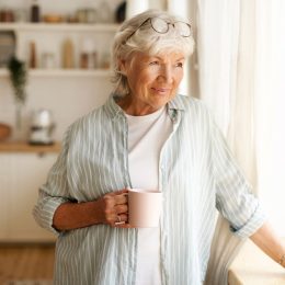 A senior woman standing by the window while holding a mug