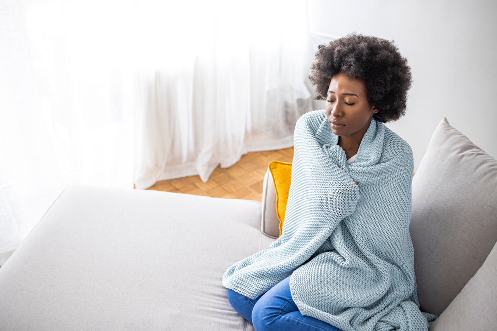A young woman with COVID-19 symptoms sitting on a couch while wrapped in a blanket