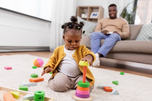 Băiețelul se joacă cu blocuri în timp ce tatăl ei se uită în fundal