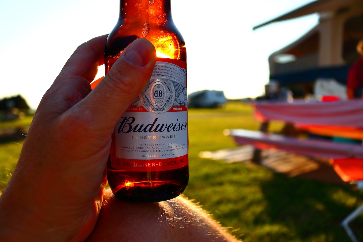 A person sitting at a campsite enjoying a Budweiser beer as the sun sets and the light highlights the bottle.