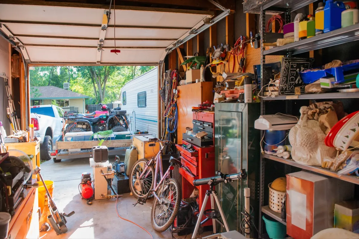 Bức ảnh này chụp một nhà để xe chứa đầy những kệ chứa đầy đồ gia dụng bao gồm dụng cụ, dụng cụ vệ sinh, đồ trang trí ngày lễ và dụng cụ thể thao.  Cửa gara đang mở.