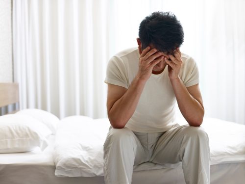 man painful and miserable sitting on bed head down covering face with hands.