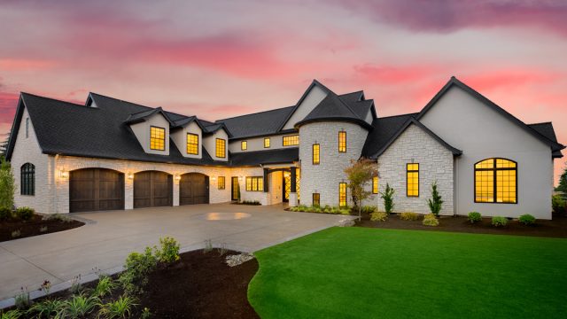 mansion with green yard and three car garage with glowing interior lights and sunset sky
