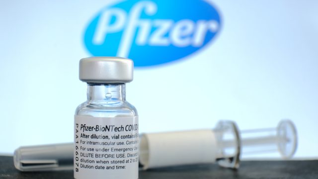 A vial of Pfizer COVID-19 vaccine and a syringe sitting in front of the company's logo