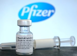 A vial of Pfizer COVID-19 vaccine and a syringe sitting in front of the company's logo