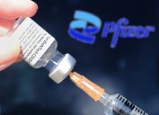 A close up of a syringe being filled with the Pfizer COVID-19 vaccine from a vial in front of the company's logo