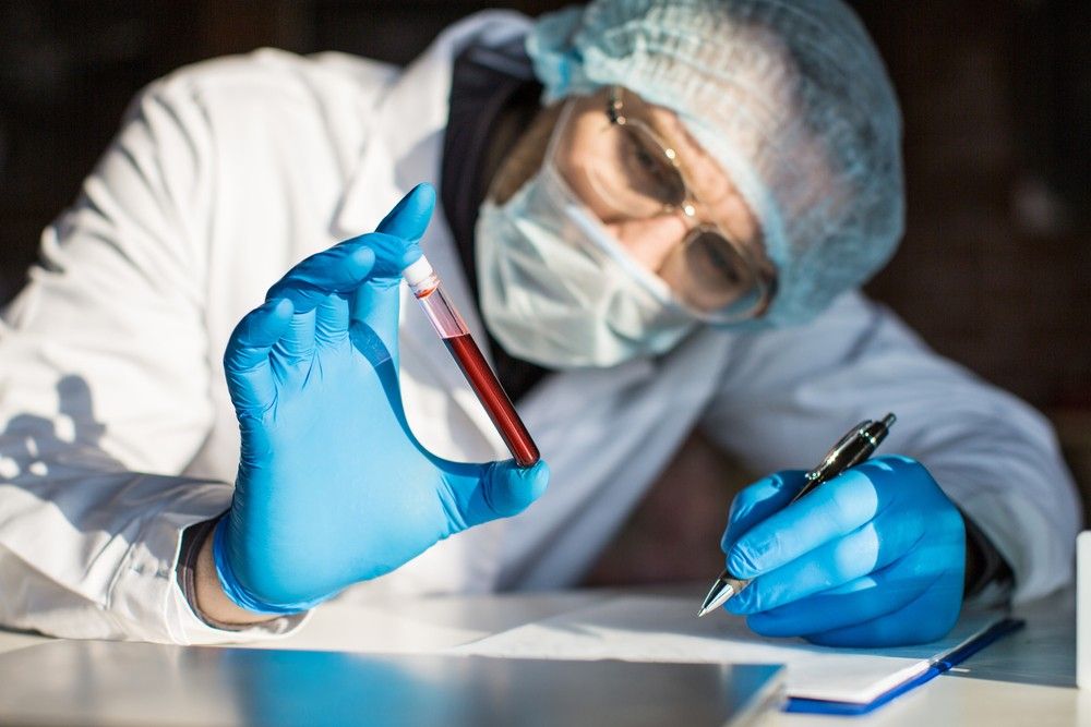 A scientist wearing full protective gear holds a vial of a blood sample while taking notes