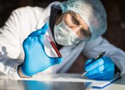 A scientist wearing full protective gear holds a vial of a blood sample while taking notes
