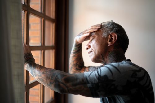 Worried mature man standing at home, close to a window, with hand in the head