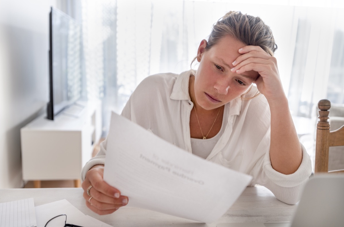 Woman looking worried holding paperwork at home. She is reading a financial bill or a letter with bad news. She looks very stressed and upset. There is a laptop computer on the table