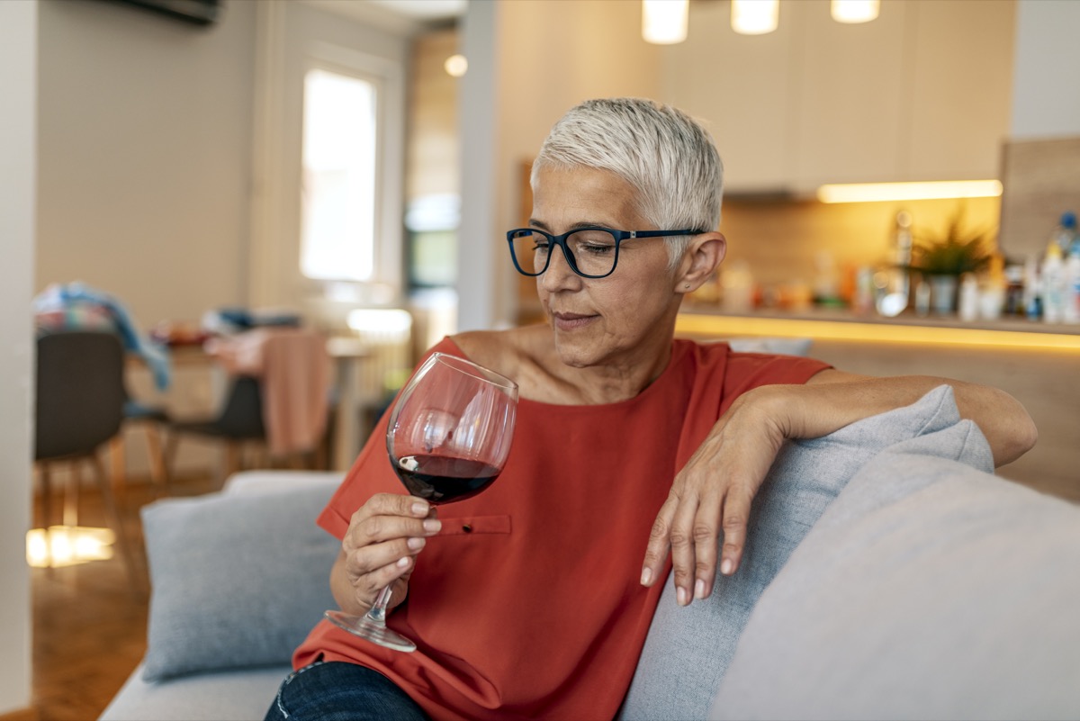 Lonely mature woman holding glass of alcoholic drink while sitting on sofa at home during the day.