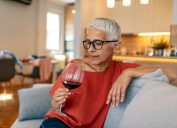 Lonely mature woman holding glass of alcoholic drink while sitting on sofa at home during the day.