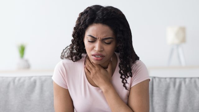 A young woman sitting on the couch and holding her throat while suffering from COVID symptoms