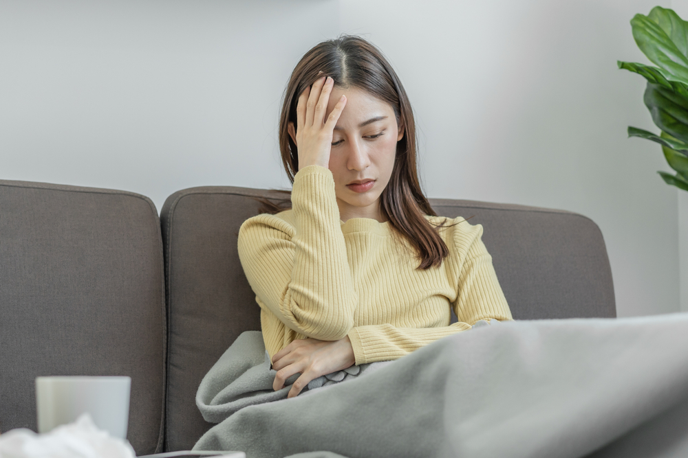 Junge Frau, die auf dem Sofa sitzt und ihren Kopf mit COVID-Symptomen krank hält