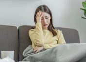 A young woman sitting on the couch holding her head while sick with COVID symptoms