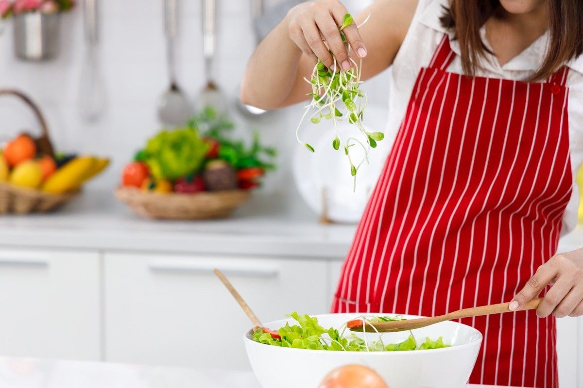 Woman chef in red apron putting sunflower sprout in salad bowl.