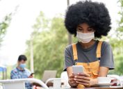 A young woman sitting outdoors at a cafe while checking her phone and wearing a mask