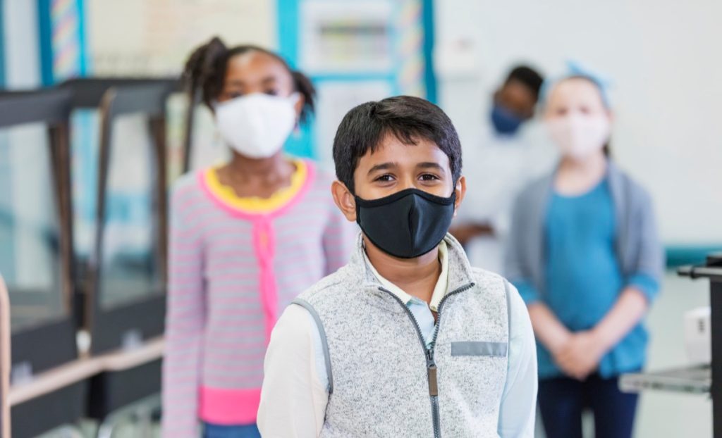 group of elementary school students in the classroom during the covid-19 pandemic, wearing face masks and standing six feet apart, social distancing.