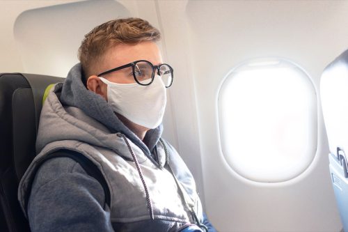 A serious guy, a young man on a plane, a plane in glasses and a sterile medical protective mask on his face on a trip.  Coronavirus, virus, airline concept.  Covid-19 pandemic.  Safety in public transport