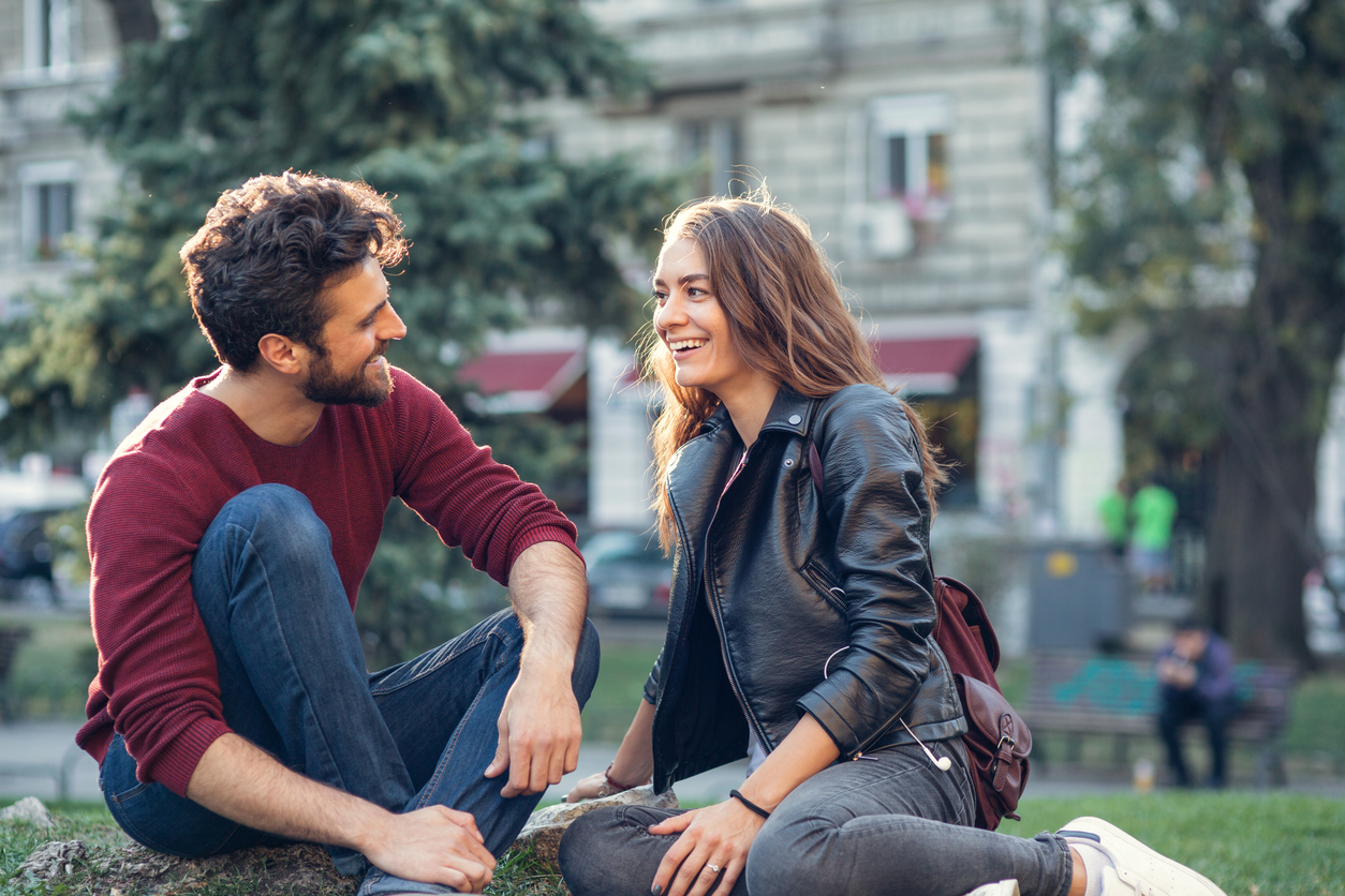 Млада двойка на среща, седнала в парк, разговаряща помежду си и усмихната