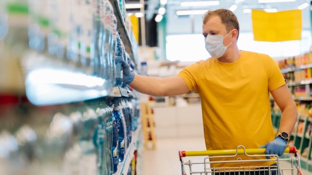Adult man wears disposable mask and rubber gloves as preventive measures during epidemic time, chooses water in retail store, tries be safe during coronavirus pneumonia outbreak. Self isolation