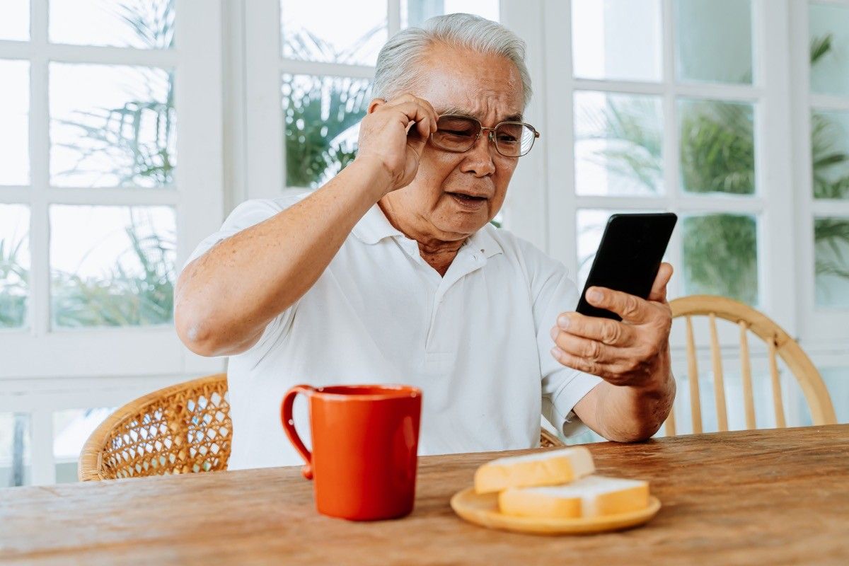 man with sight problem and wearing glasses while watching and reading message on smartphone during breakfast at home.  Old man with blurred eyes vision indoors.