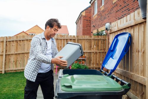 Side view of a senior man disposing of his household recycling into an outdoor bin in his garden. He is in the North East of England.
