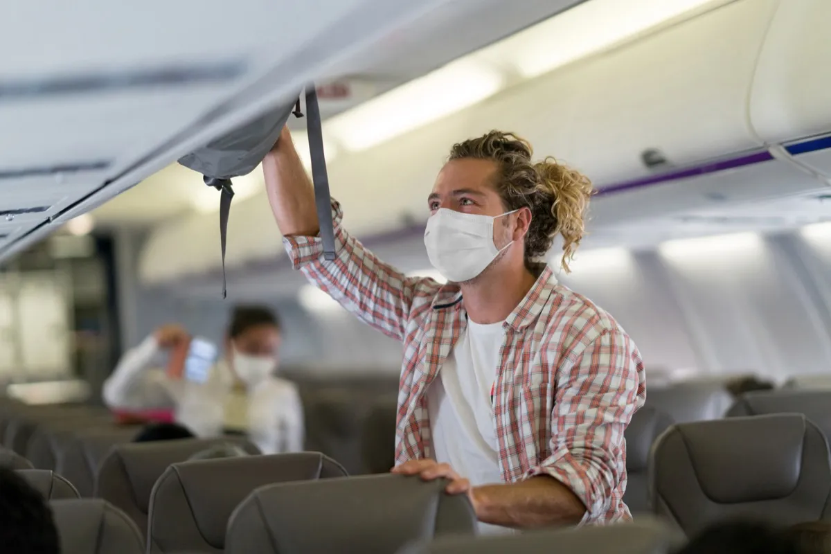 Passenger putting his carry-on luggage in the overhead compartment and wearing a facemask inside the airplane during the COVID-19 pandemic