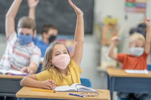 Group of students wearing protective face masks while raising their hands in class.
