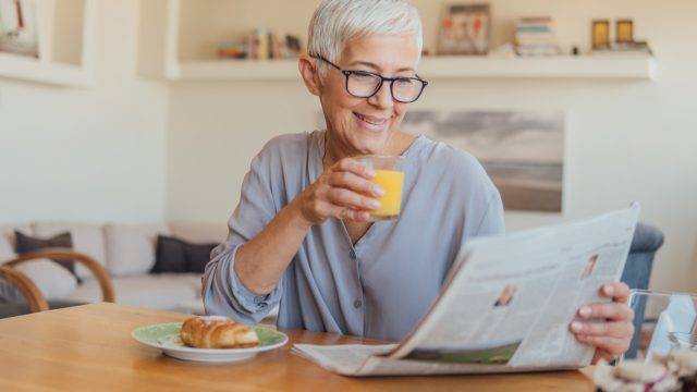 A senior woman drinking juice at breakfast while reading the paper