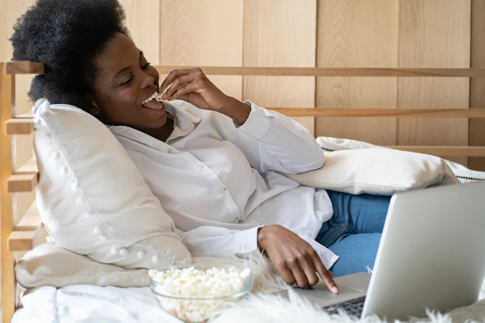 A young woman sitting in bed while watching a streaming service on her laptop and eating popcorn