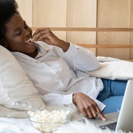 A young woman sitting in bed while watching a streaming service on her laptop and eating popcorn