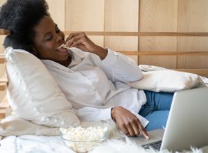 A young woman sitting in bed while watching a streaming service on her laptop and eating popcorn