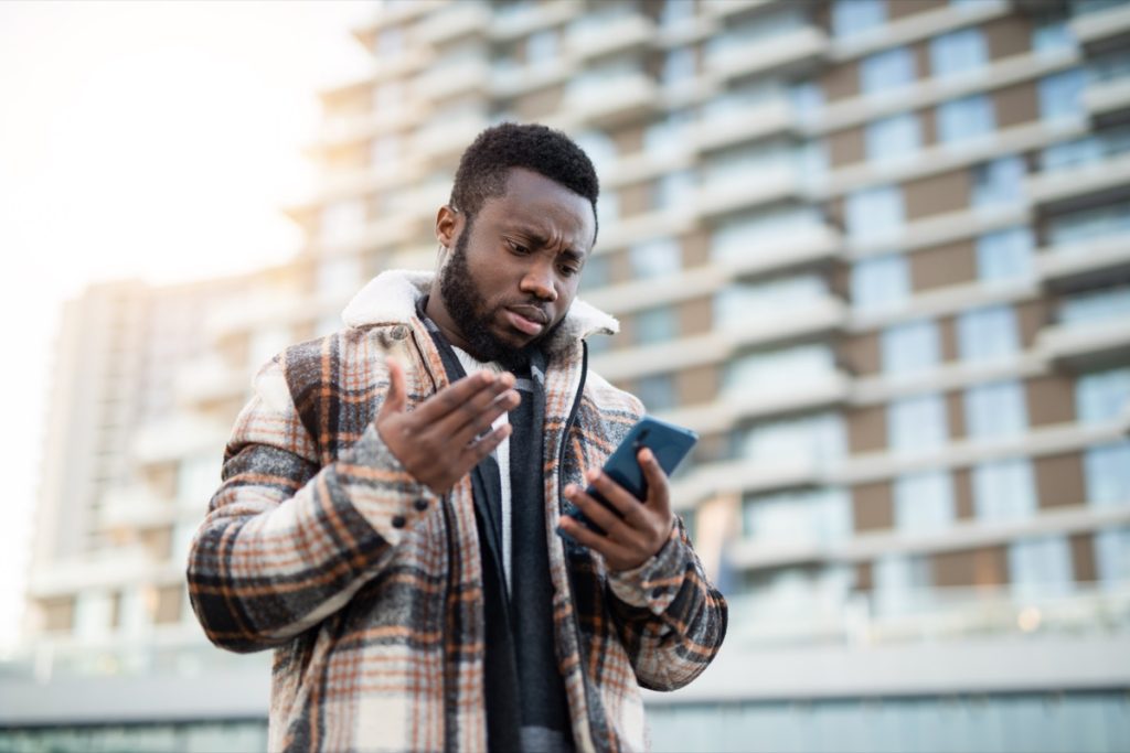 man standing outside apartment building looking frustrated at phone