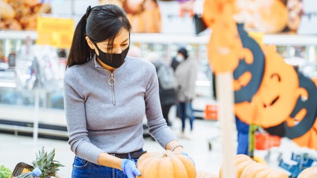 Young woman wearing a protective mask and gloves choosing pumpkins for Halloween in a supermarket. Reality 2020.
